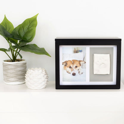 Black photo frame with a dog portrait and paw print on a white surface with a plant and decorative item.